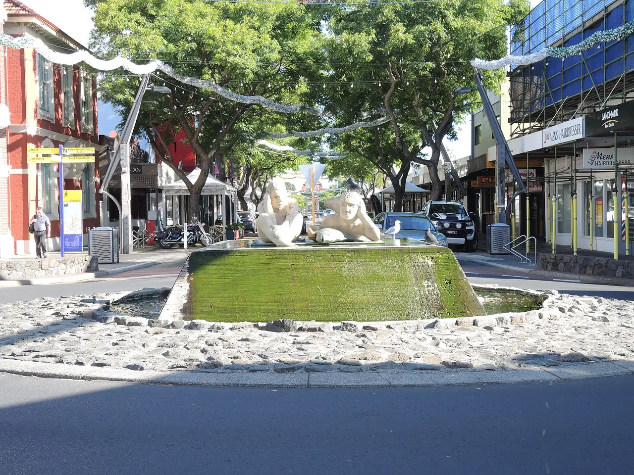 A white sculpture and a fountain on a tree-lined shopping street