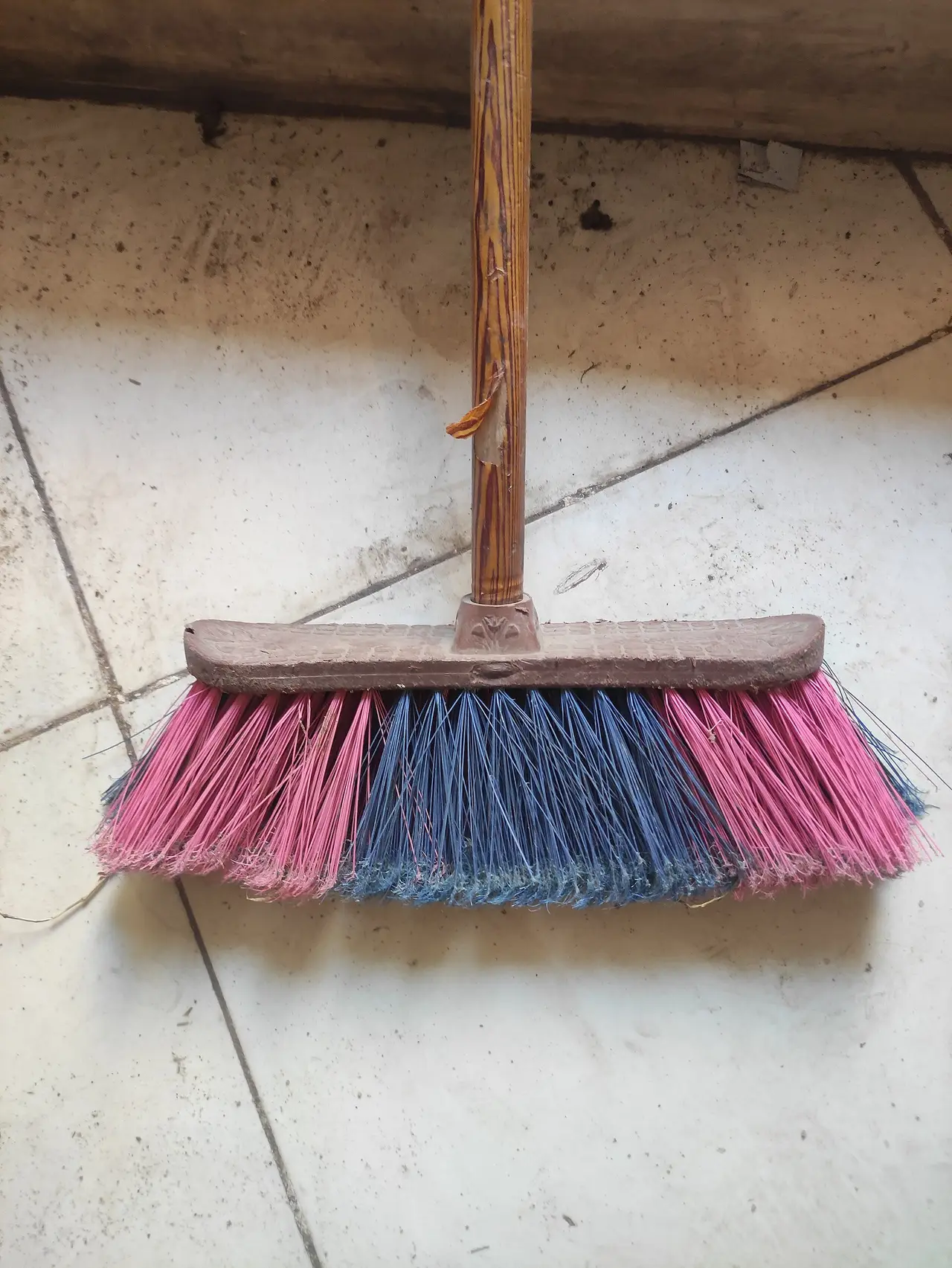A broom with pink and blue bristles resting on a tiled floor