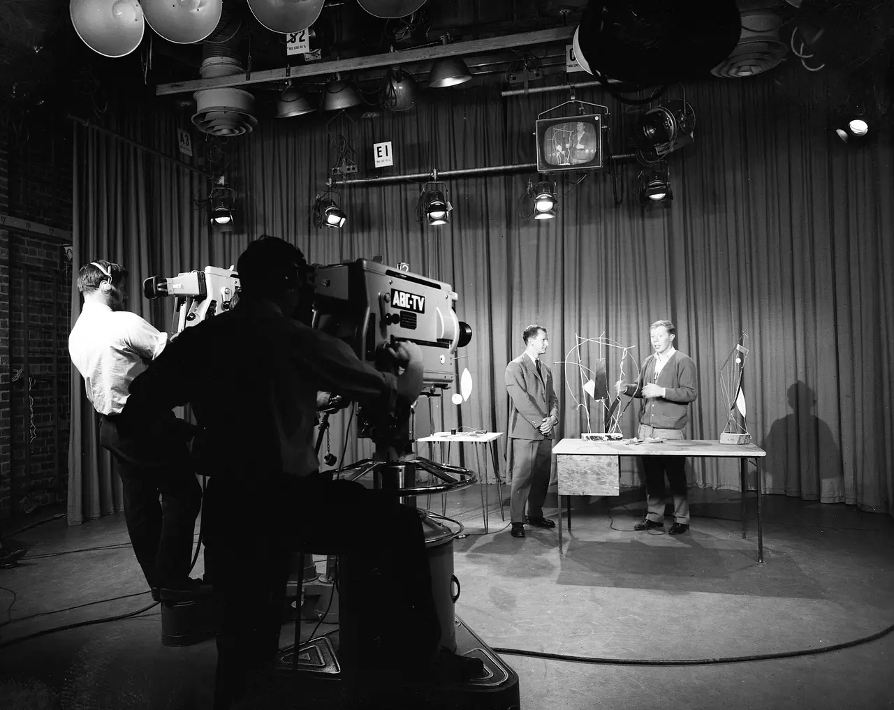 Black-and-white view of a television studio with a camera and two men at a desk