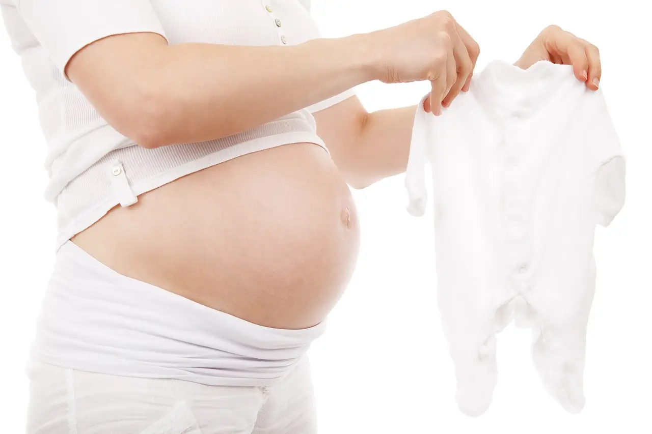 A pregnant woman holding a white baby sleepsuit next to her bump
