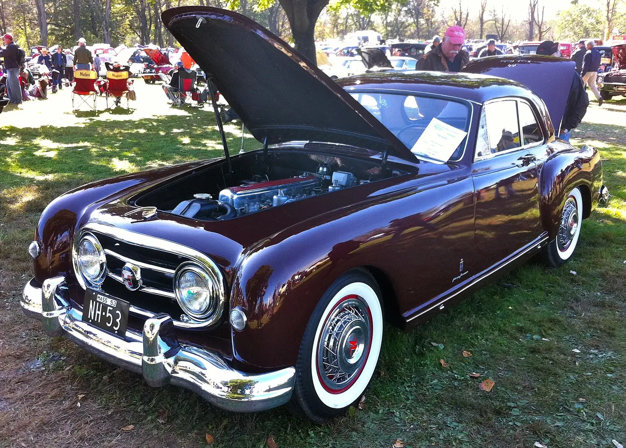 A dark red 1950s coupe with its bonnet open at a car show