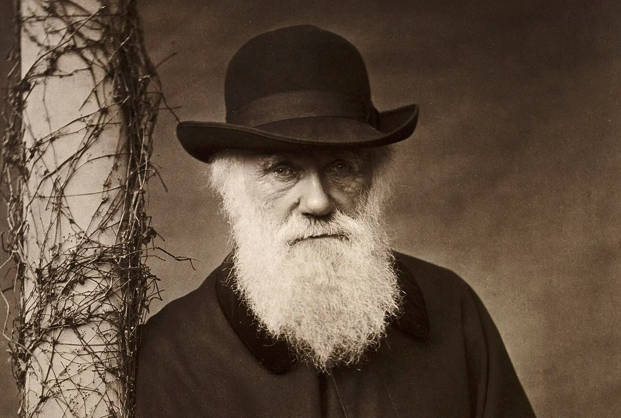 Charles Darwin with a white beard and a brimmed hat beside a vine