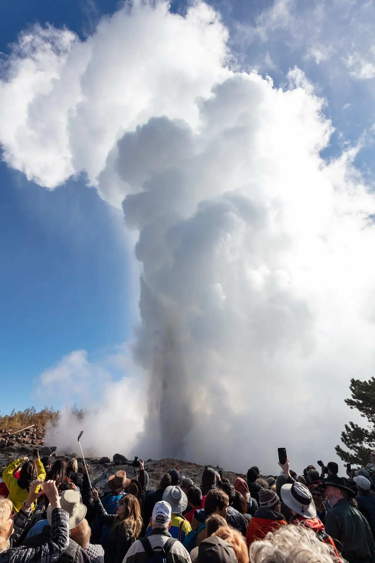 A crowd with phones raised watching a geyser throw steam into the sky