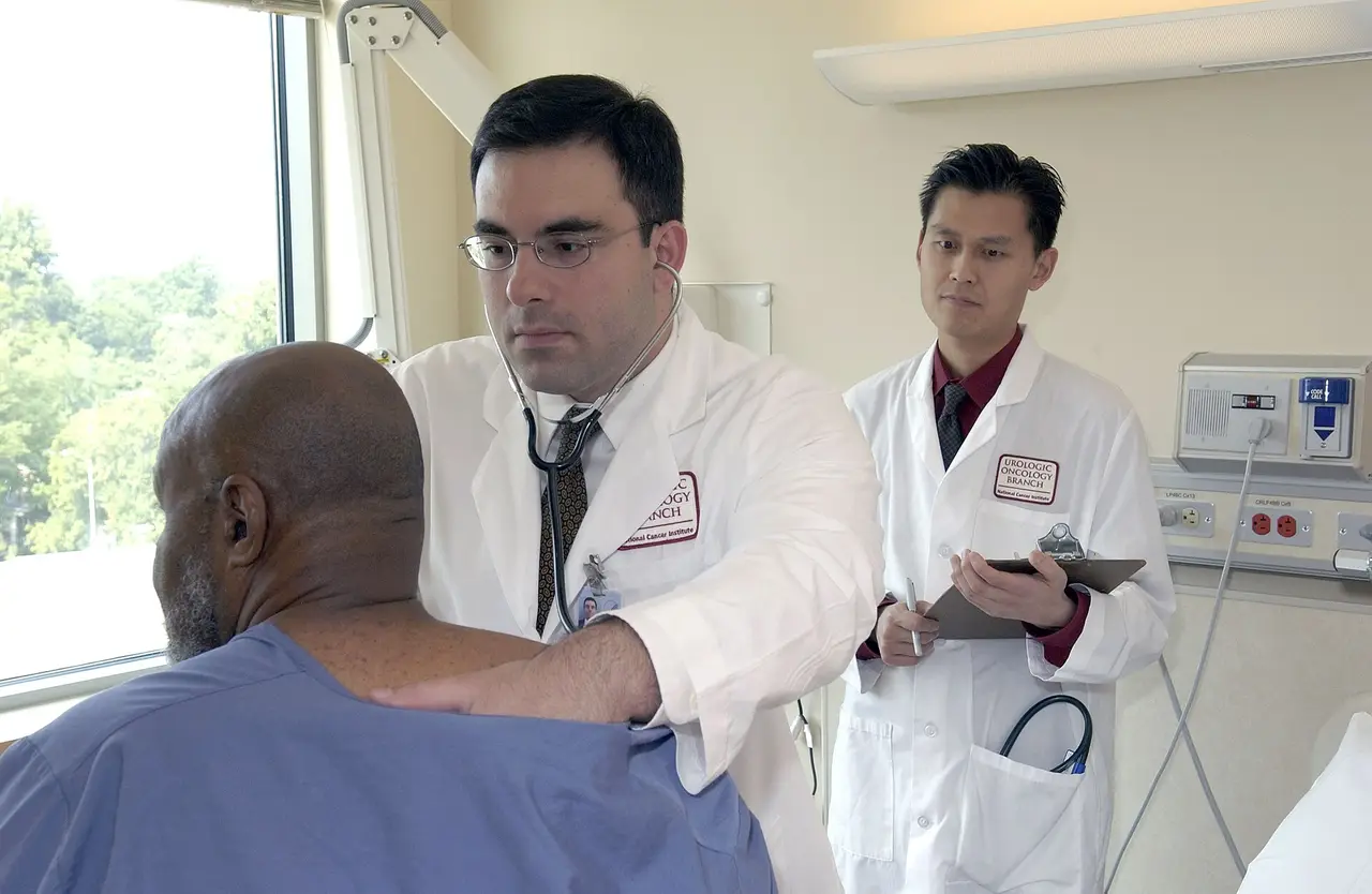 A doctor in a white coat listens to a seated patient's chest with a stethoscope