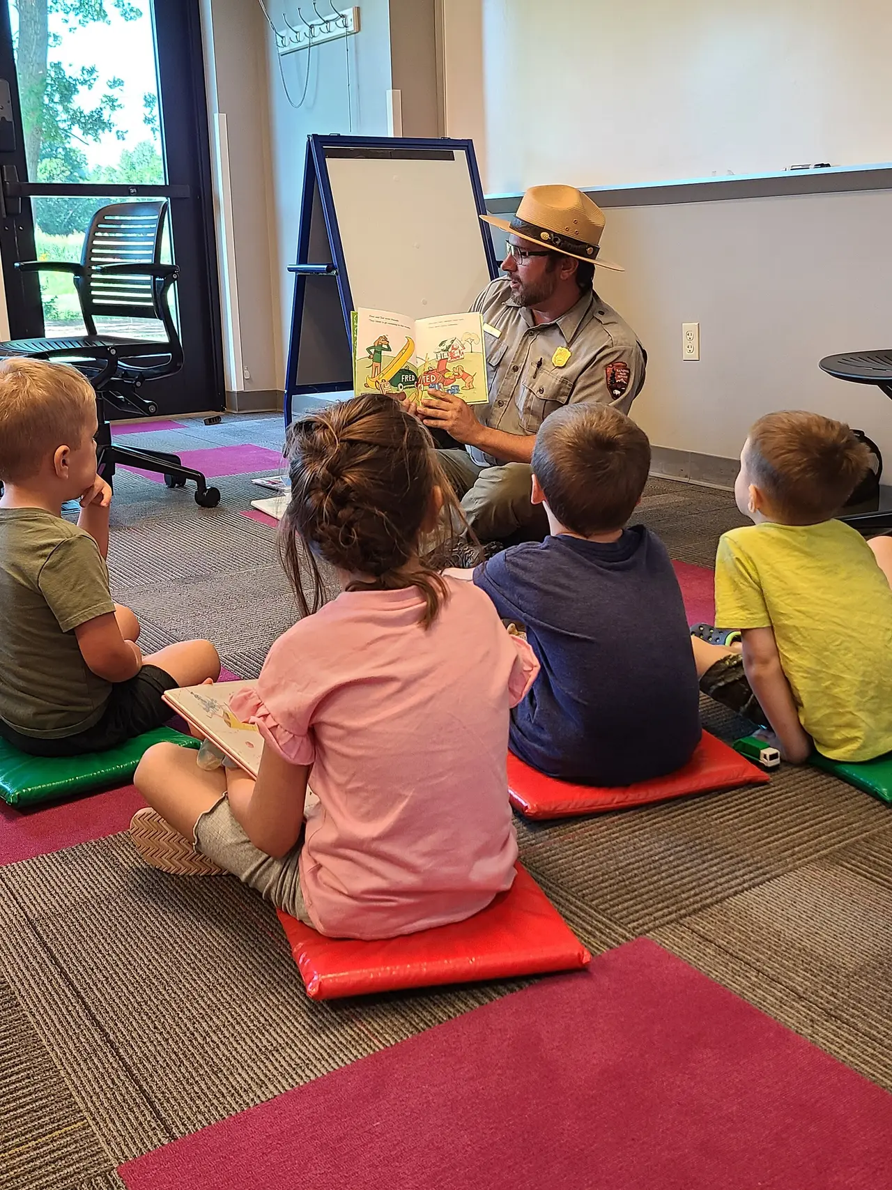 A ranger holding a book sits in front of children on floor mats
