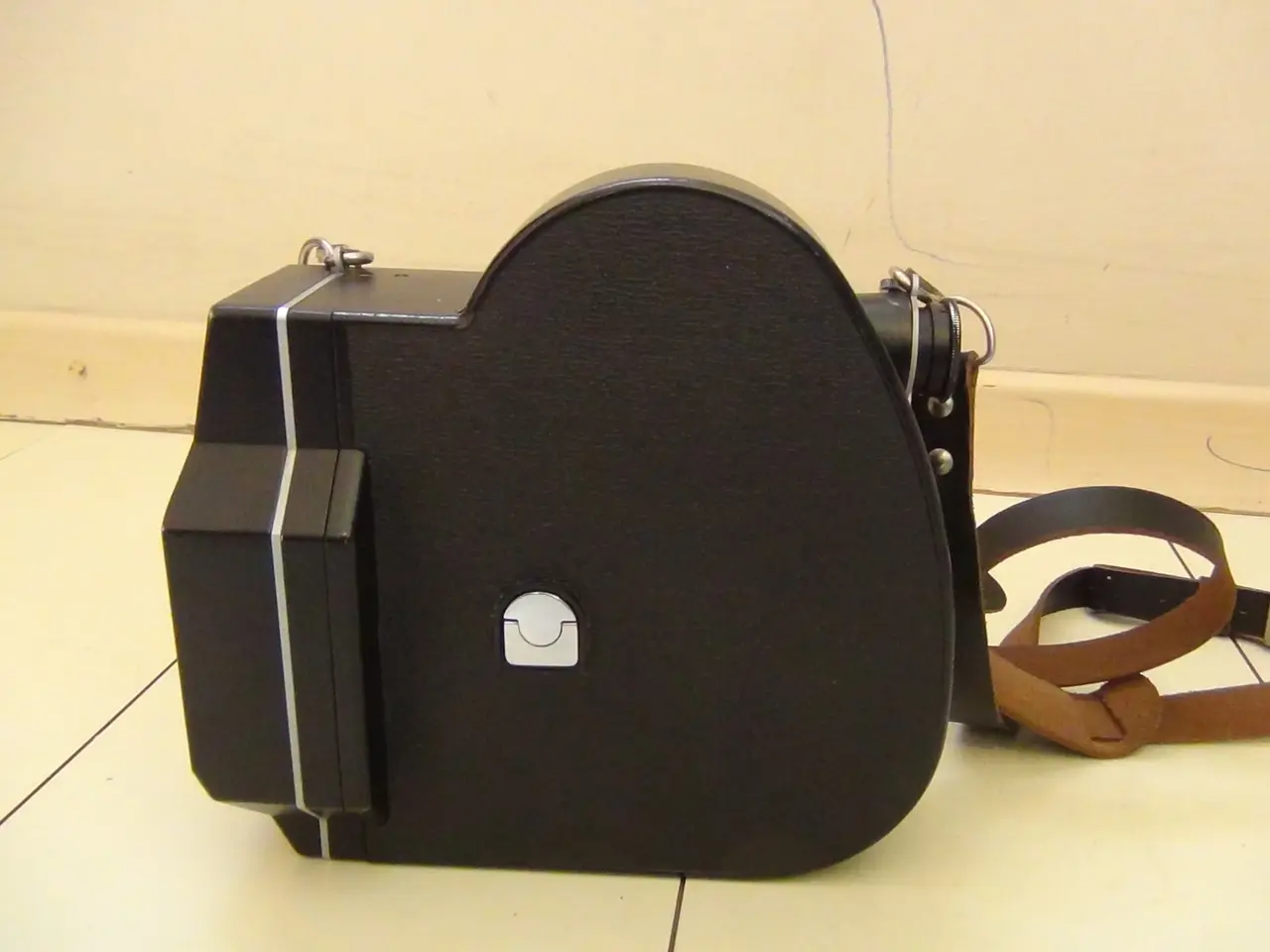 A black wind-up movie camera with a brown strap on a tiled floor