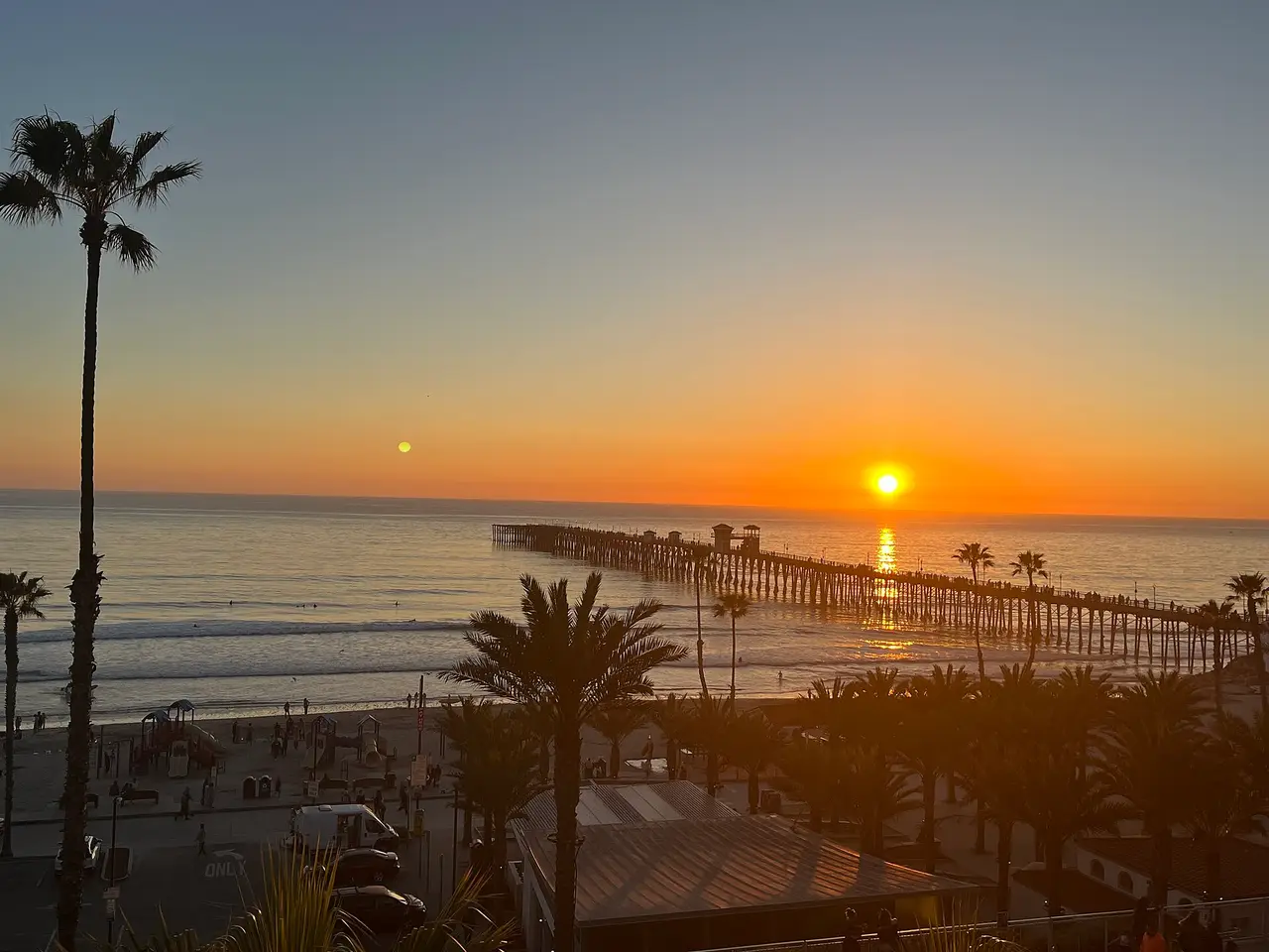The sun setting over the sea beside a long pier and a palm tree