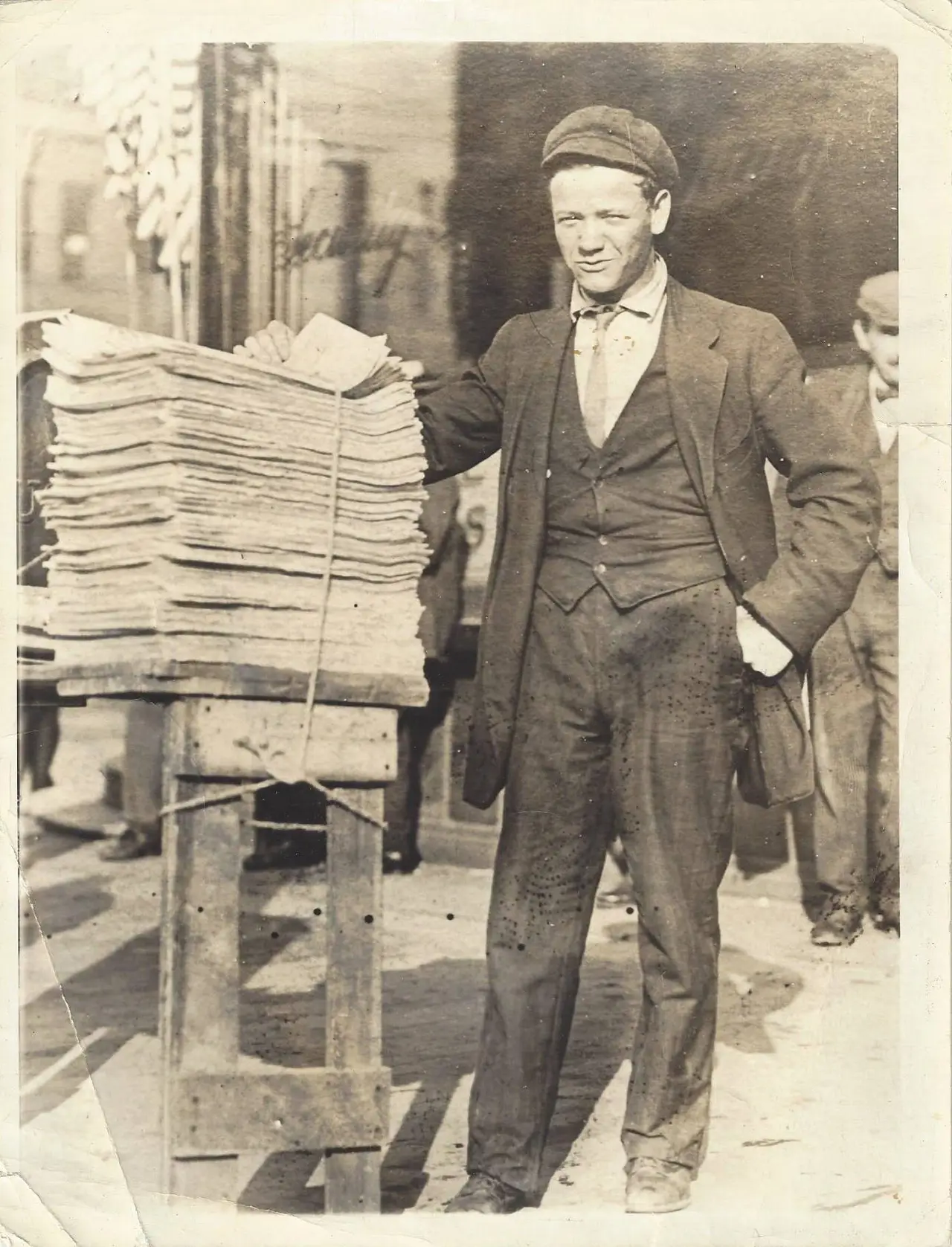 A man in a waistcoat and cap beside a stack of newspapers