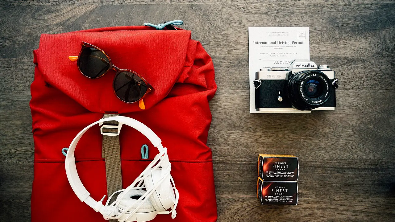 A red backpack beside sunglasses, a camera and headphones