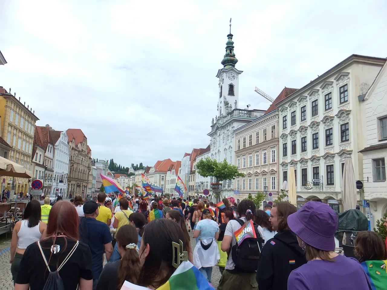 A crowd with rainbow flags walking along a town square lined with buildings