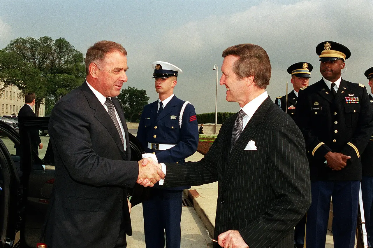 Two men in dark suits shaking hands in front of uniformed servicemen