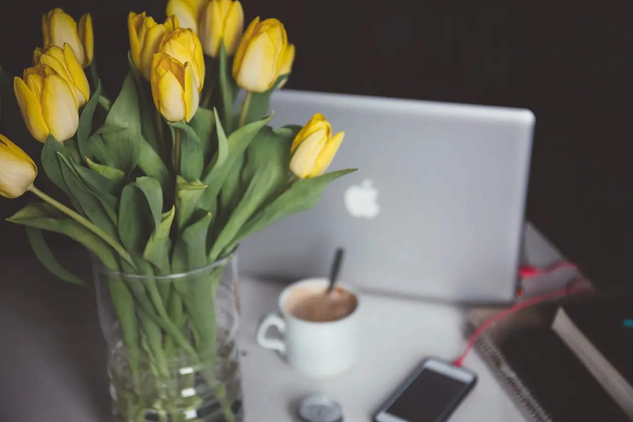 Yellow tulips in a glass vase beside a laptop and a coffee cup
