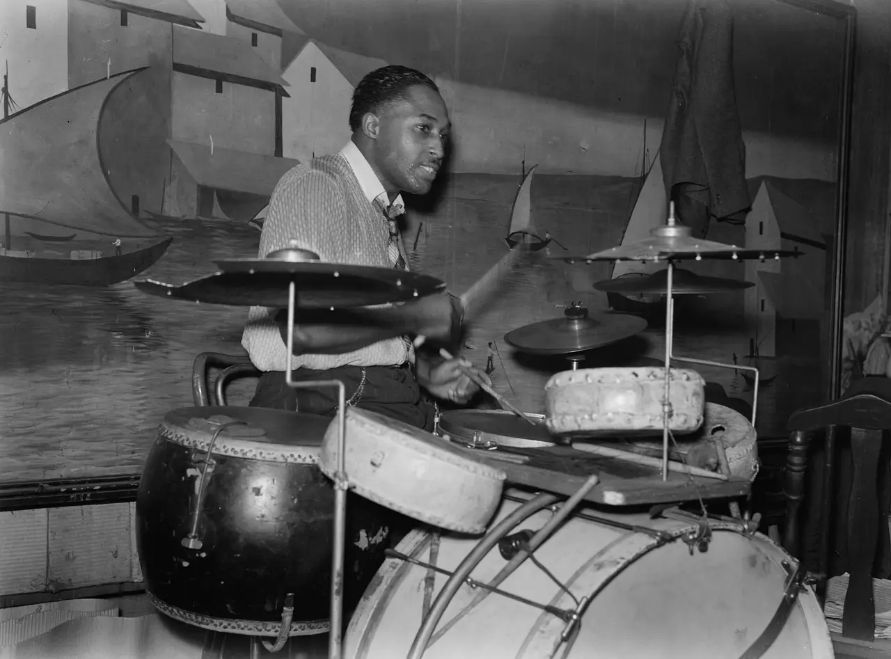 A drummer seated behind a bass drum and cymbals in a black and white photograph