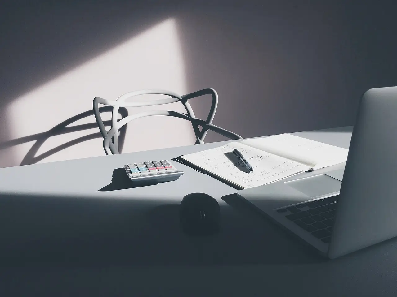 A desk with a laptop, calculator, notebook and pen in window light