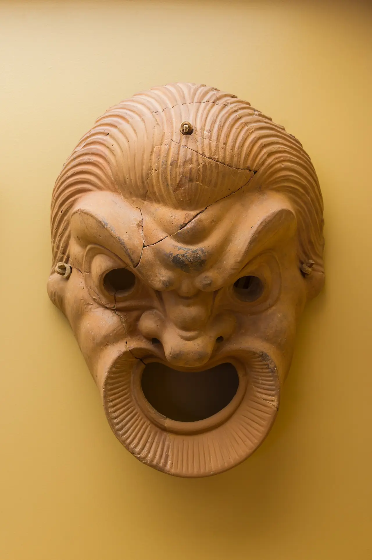 A clay theatre mask with grimacing face and open mouth on a yellow wall