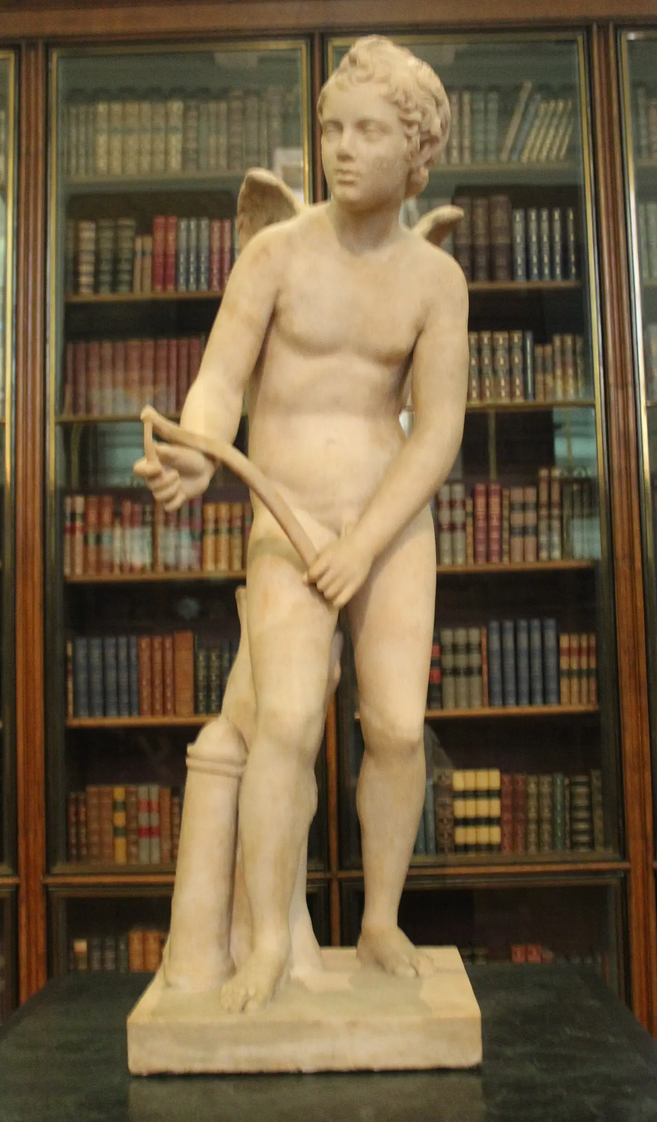 A pale marble statue of Cupid with wings standing in front of bookshelves