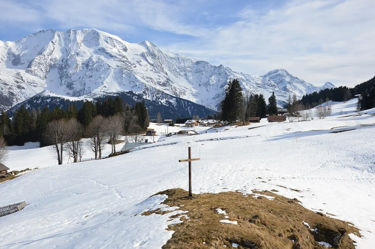 A small wooden cross on a mound in a snowy field below mountains