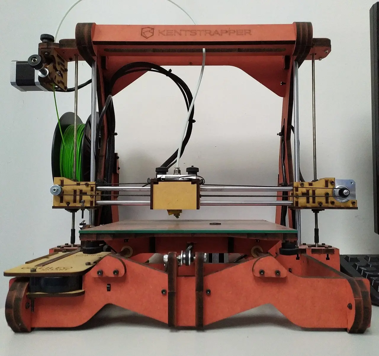 A red wooden-framed 3D printer with a spool of green filament and loose wires