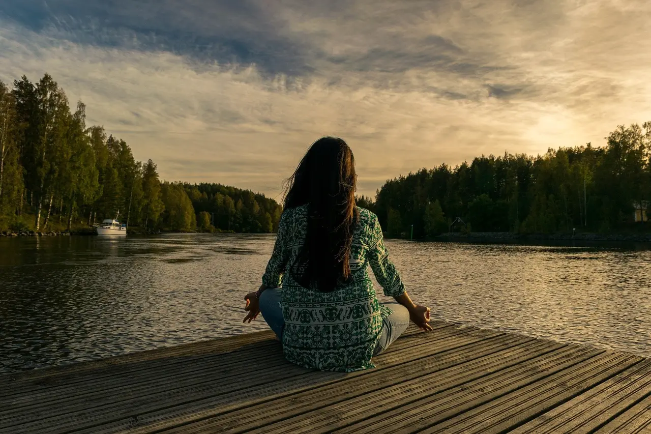 A woman sitting cross-legged on a jetty facing a lake at sunset