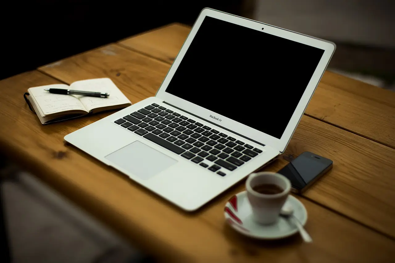 An open laptop with a notebook, phone and coffee cup on a wooden desk