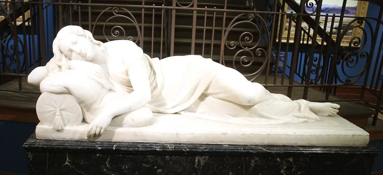 A white marble statue of a girl asleep on a bolster, on a black plinth