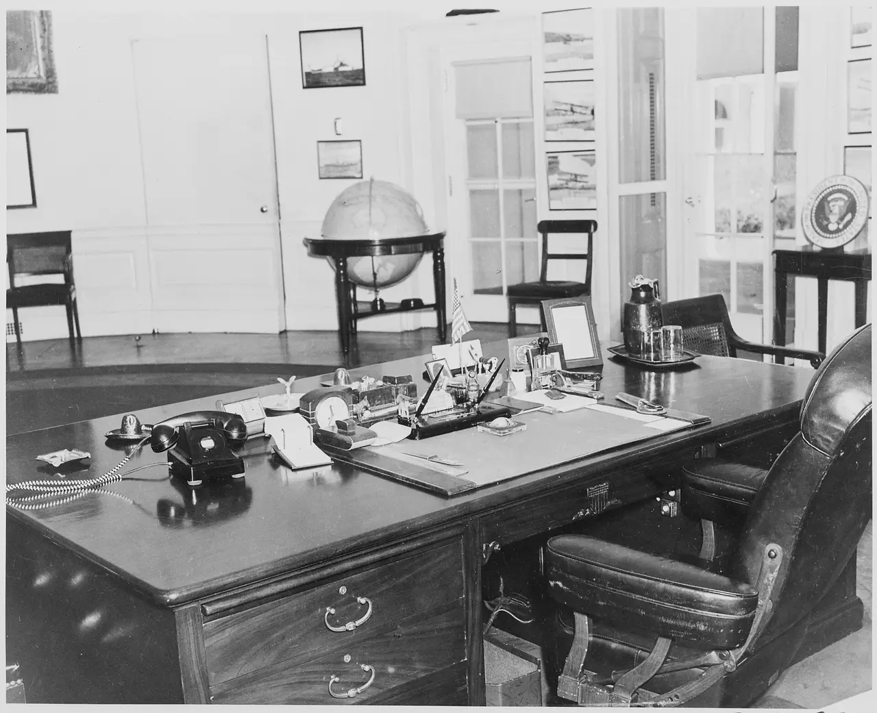 A black and white view of a wooden desk with a telephone, papers and a globe behind