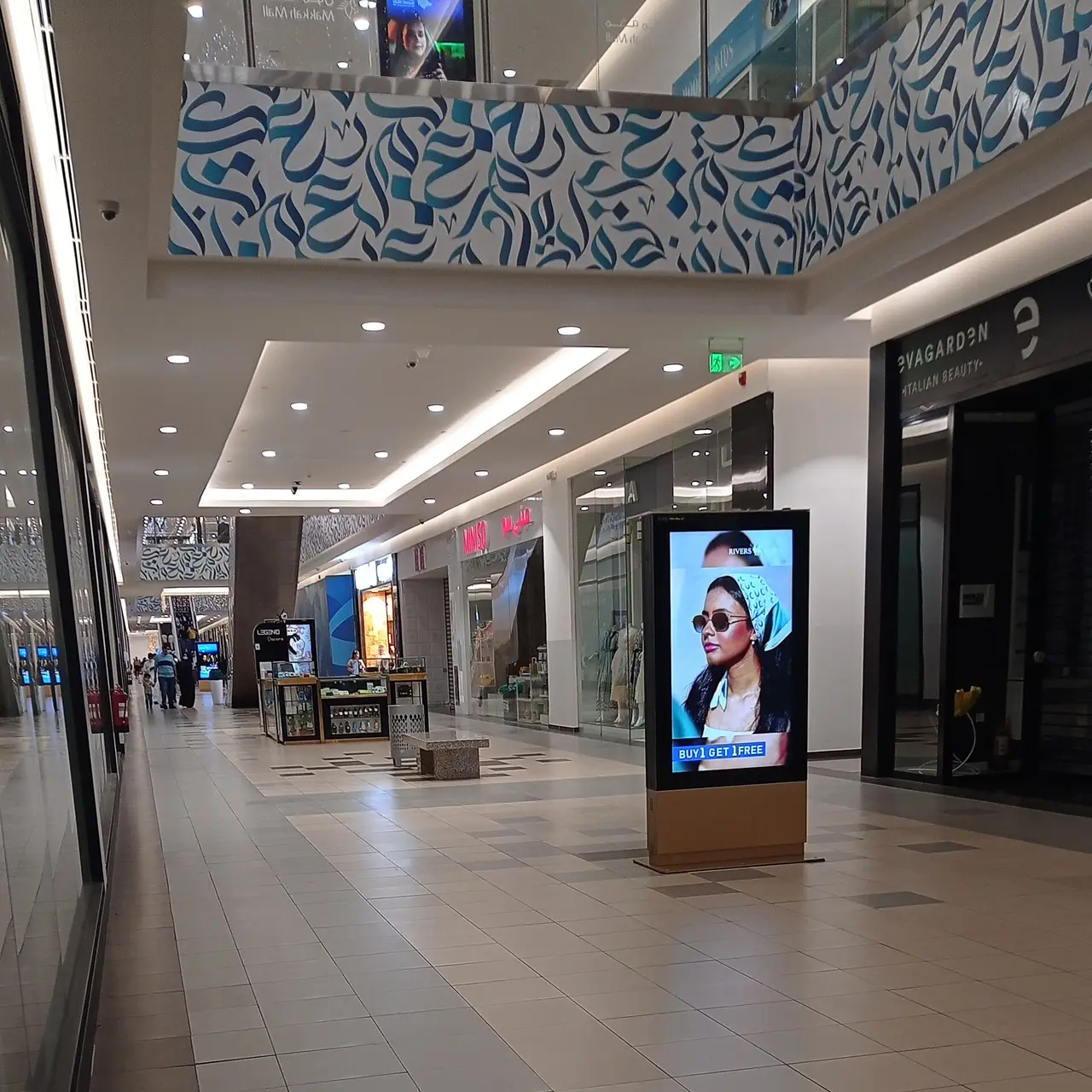 A long shopping mall passage with shop fronts and a lit advertising screen