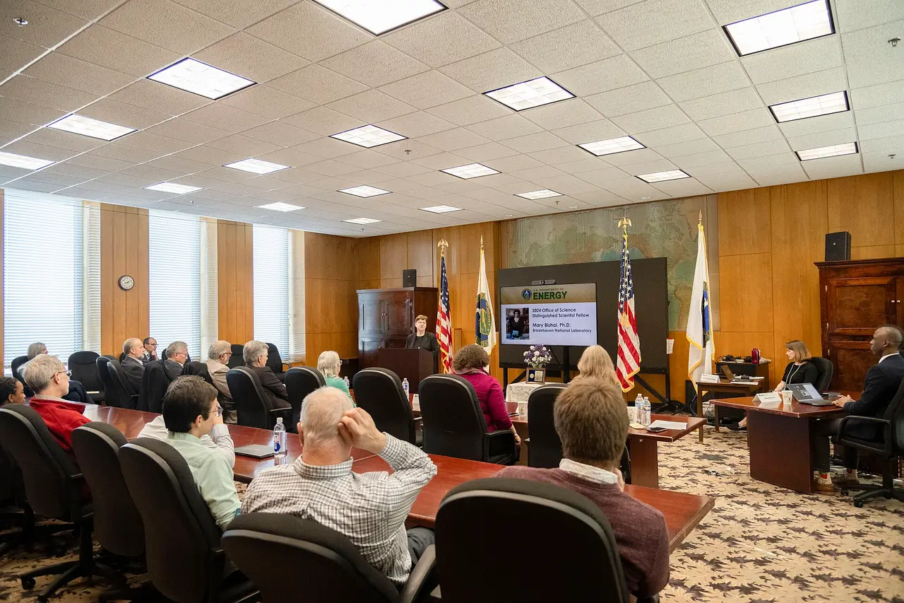 Seated people at long tables facing a screen and lectern in a meeting room