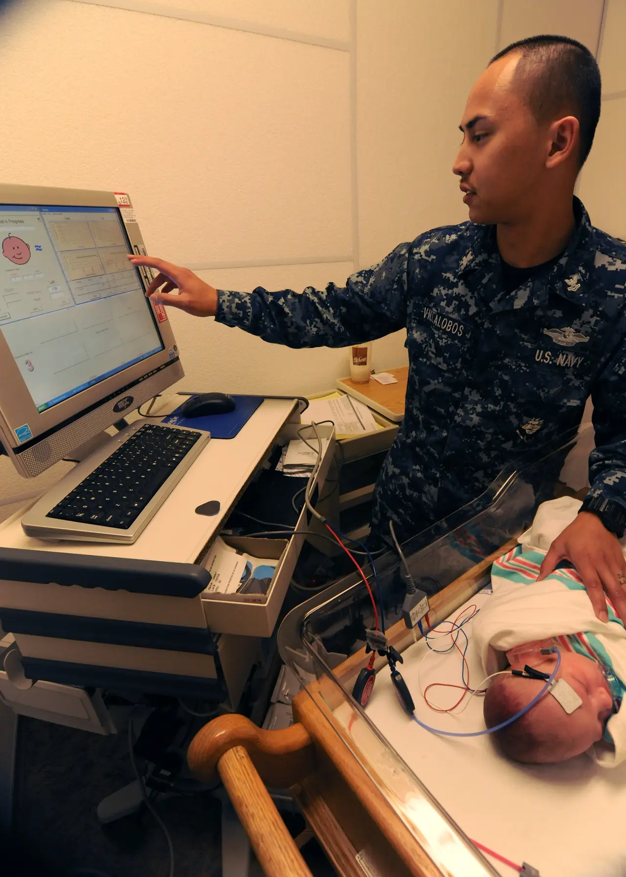 A uniformed corpsman looks at a monitor beside a newborn screening setup