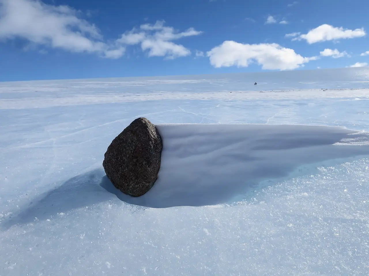 A dark rock sitting on a white and blue ice field under a clear sky