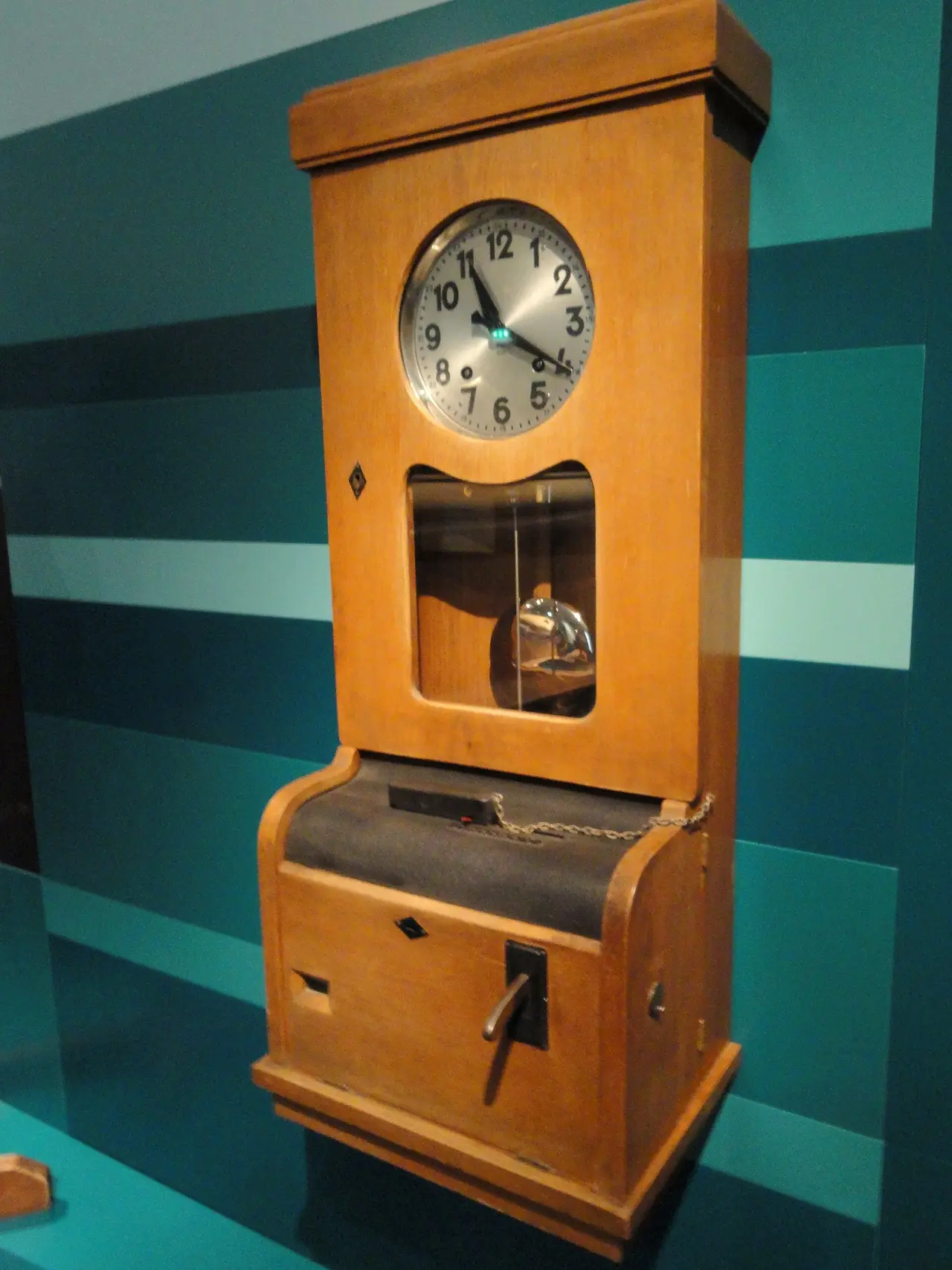 A tall wooden clock with a round dial on a teal wall