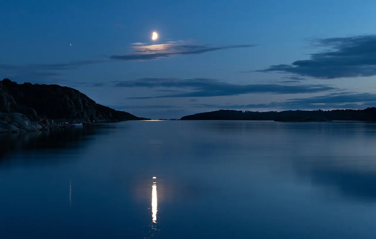 A half moon above a still fjord with its reflection on the water