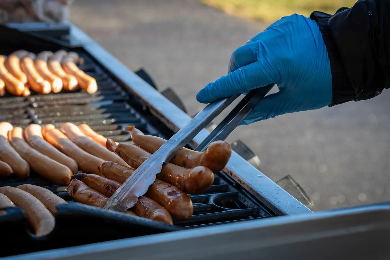 A gloved hand turning rows of hot dogs on a grill with tongs