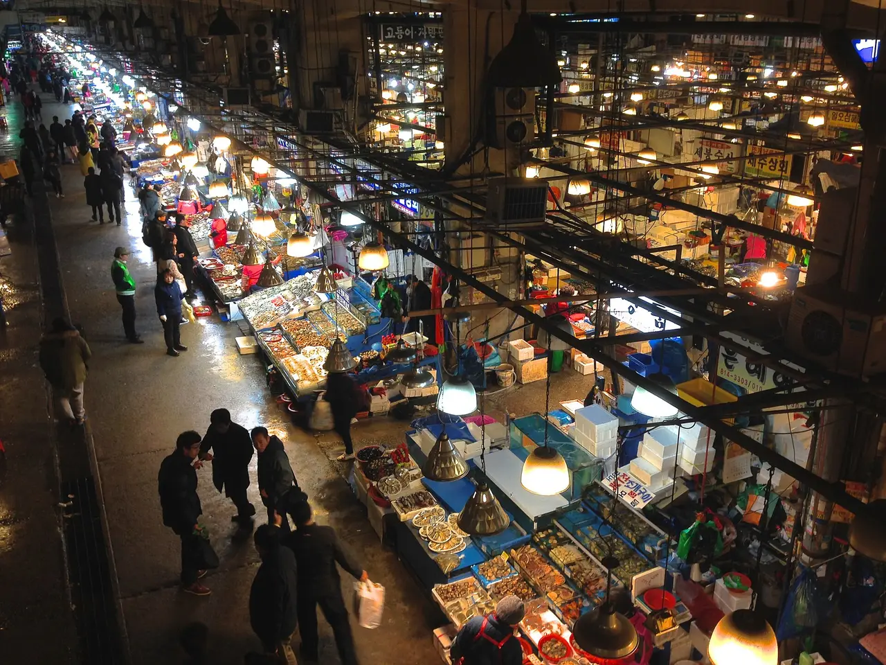 Shoppers between brightly lit seafood stalls in a covered fish market at night