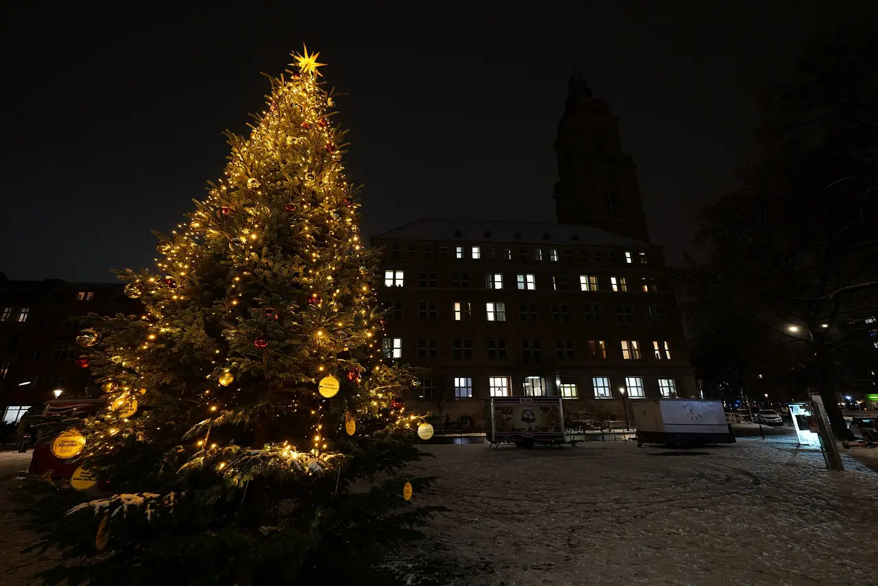 A lit Christmas tree on a snowy square at night with buildings behind