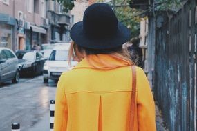 back view of young woman in yellow coat and black hat on street