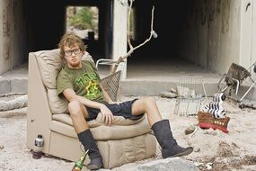 young man in glasses sits on soft chair in dirty yard