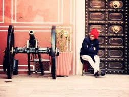 old man in traditional clothing sits at wall of fort beside of cannon, india, rajasthan, mughal