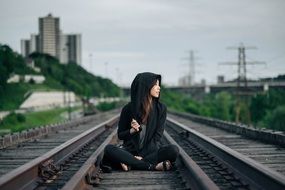 hipster, young asian girl in black clothing sits between rails on railroad