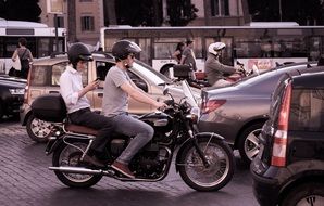 two young men in helmets on motorbike among cars on road in city