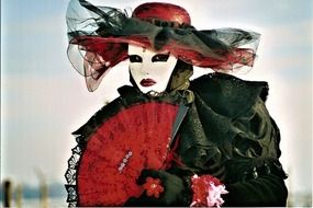 black and red dressed person in white woman mask with red fan