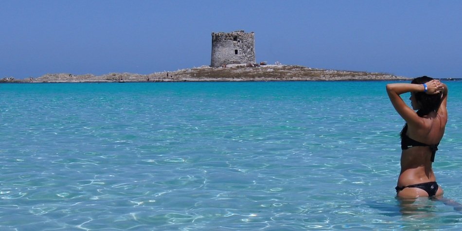 woman in bikini stands in ocean in view of old tower on island