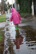 little girl at pink raincoat stays in puddle at street