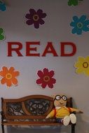 doll sits on bench at wall under red lettering read and paper flowers