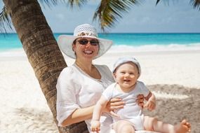 joyful woman with baby boy sits on sand beneath palm tree