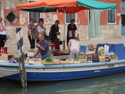 vendor in boat with vegetables on street market, italy, venice