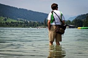 man with bag knee-deep in water at beautiful landscape with plants and mountains
