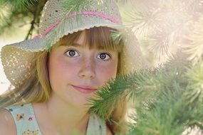 Little girl in a wide-brimmed hat among fir branches