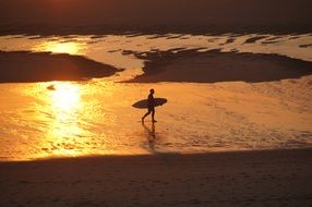 man with a surfboard is walking along the beach in the orange twilight