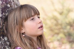 blonde girl leans on tree at elbows at blurred background with the plants